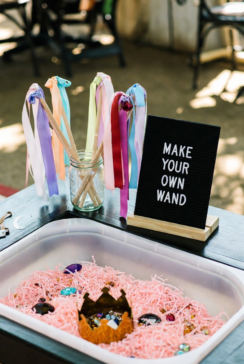 A make-your-own-wand station: ribbon wands standing in a mason jar beside a black letterboard reading MAKE YOUR OWN WAND, with a tray of pink shred and jewels.