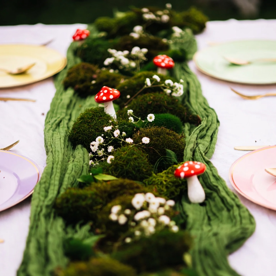 Green gauze runner topped with clumps of moss, sprigs of baby's breath, and red-and-white toadstool mushrooms, between pastel plates.