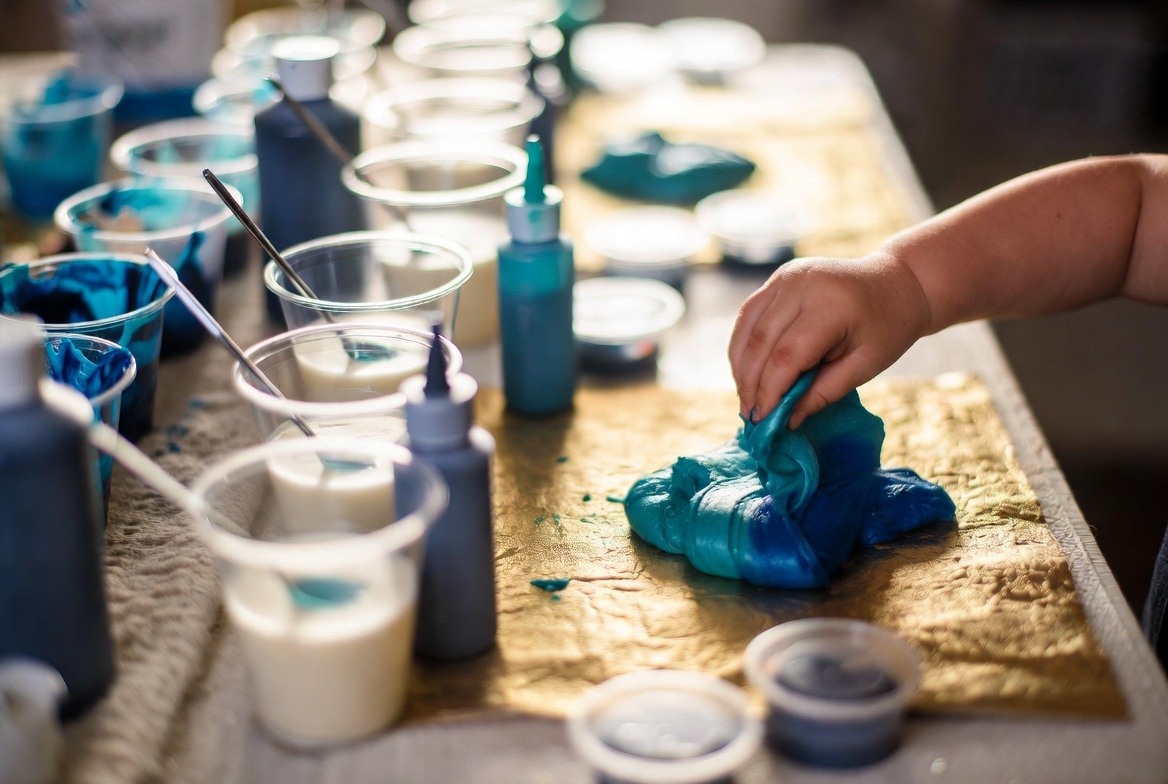 Toddler's hand mixing bright blue slime with cups of paint and bottles on a gold mat