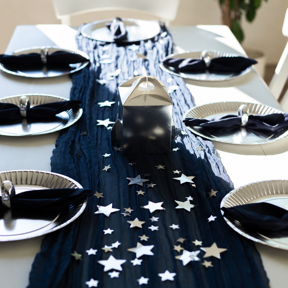 Navy cheesecloth runner on a white table scattered with silver foil stars trailing from a white treat box, with navy napkins on silver plates.
