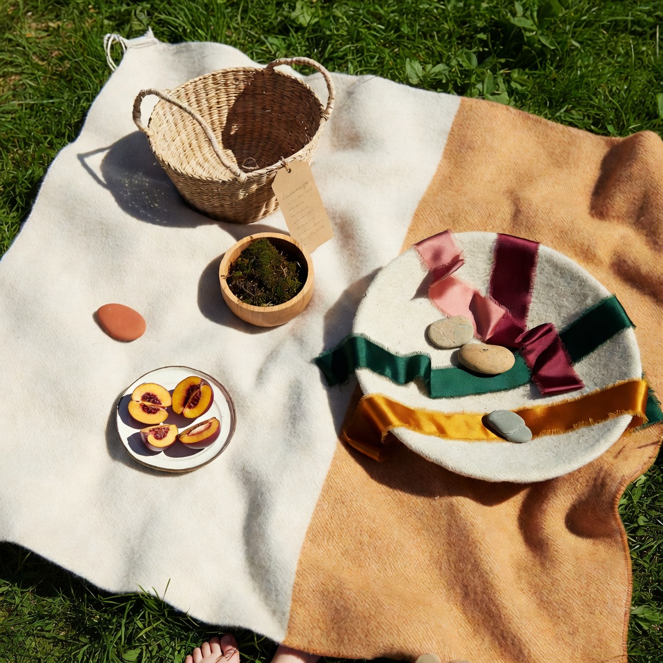 Natural wool picnic blanket on grass with a basket, fruit plate and ribbons