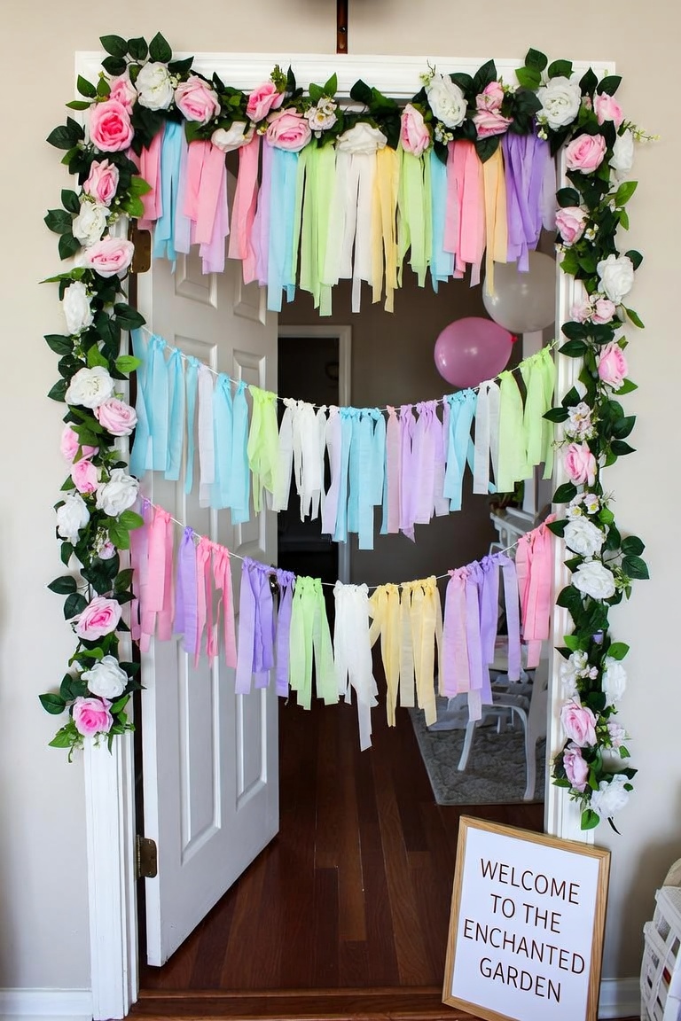 Doorway framed with faux rose-and-greenery garland and three rows of pastel fabric-strip banners, with a Welcome to the Enchanted Garden letterboard sign.